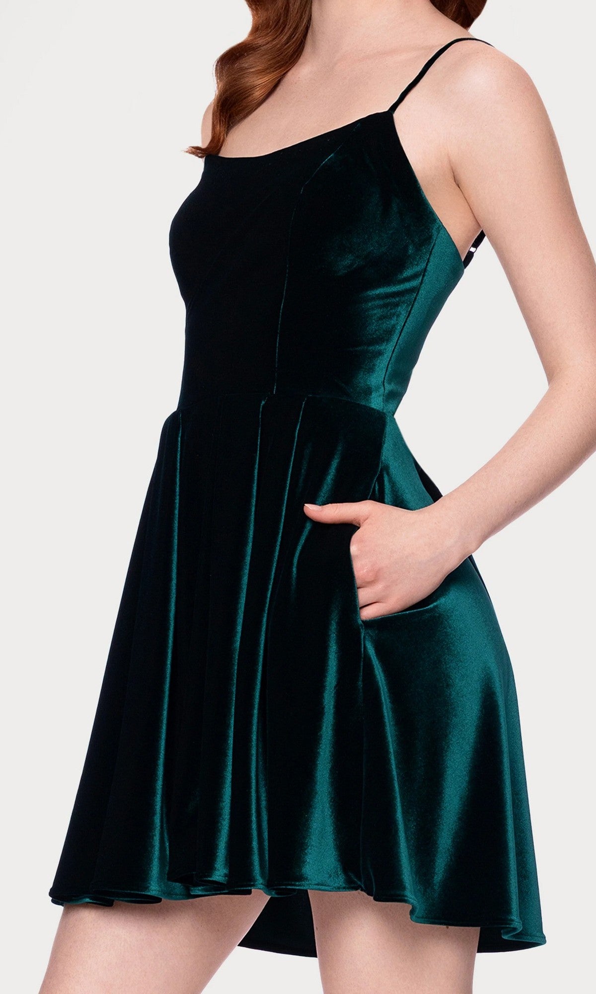 A woman wearing the Blondie Nites 5601BN short prom dress—a sleeveless, dark green velvet junior dress with thin straps and an a-line skirt—stands with one hand in her pocket.